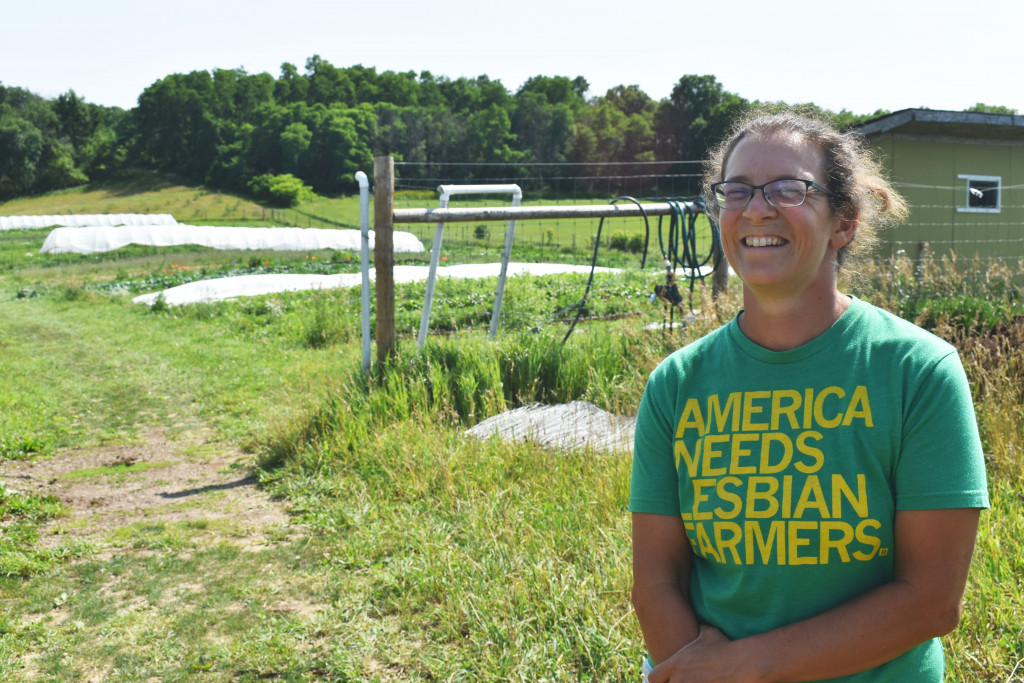 A young woman in glasses and a shirt that reads "America needs Lesbian Farmers." smiles in front of a background of agricultural fields.