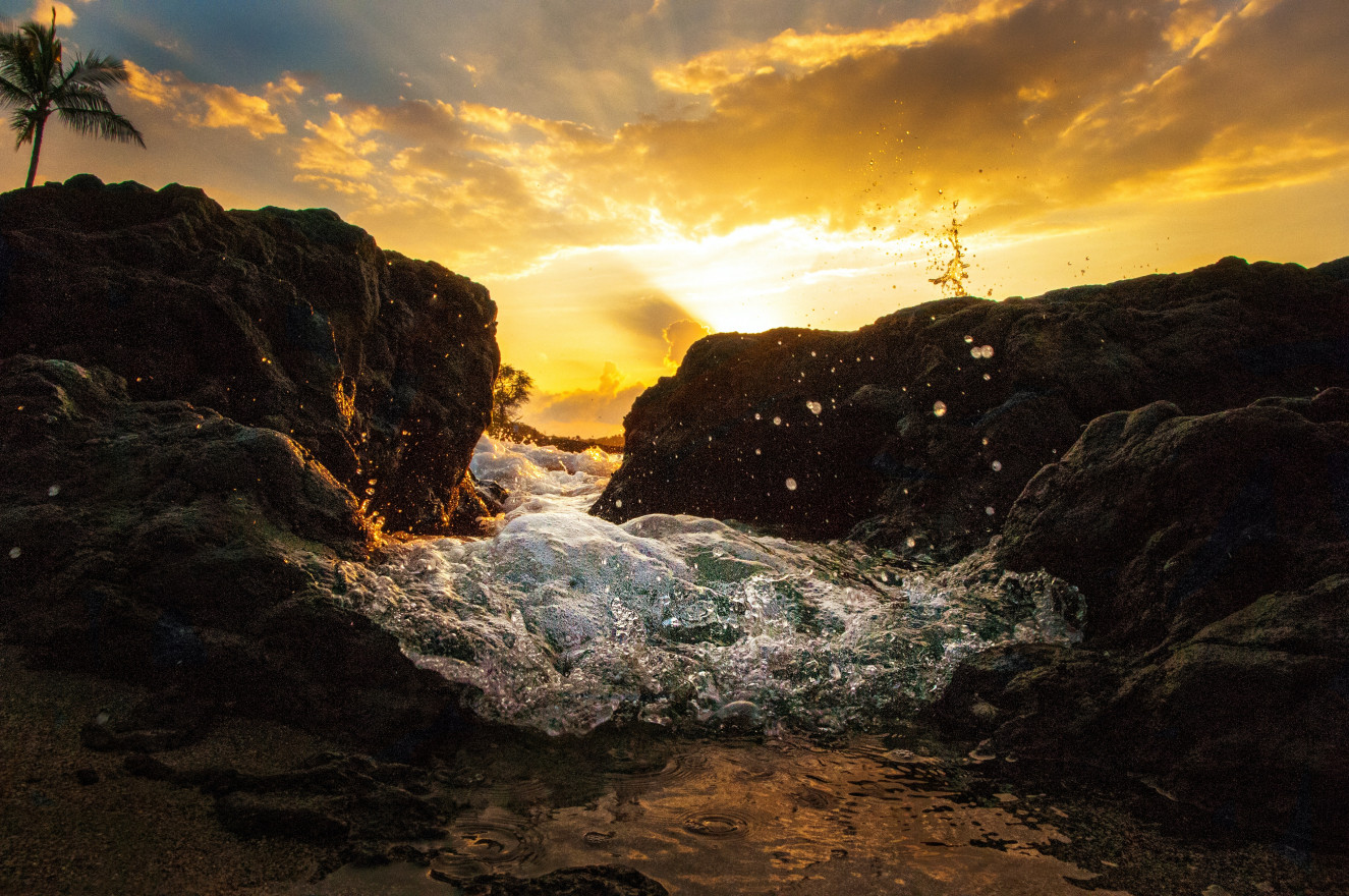 A tide splashing in between two rocks on a coast line as the sun sits low in the sky behind it.