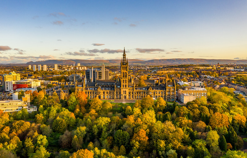 Glasgow university, with a distinctive central spire, is seen from the air, with a cluster of trees turning autumnal colors in the foreground