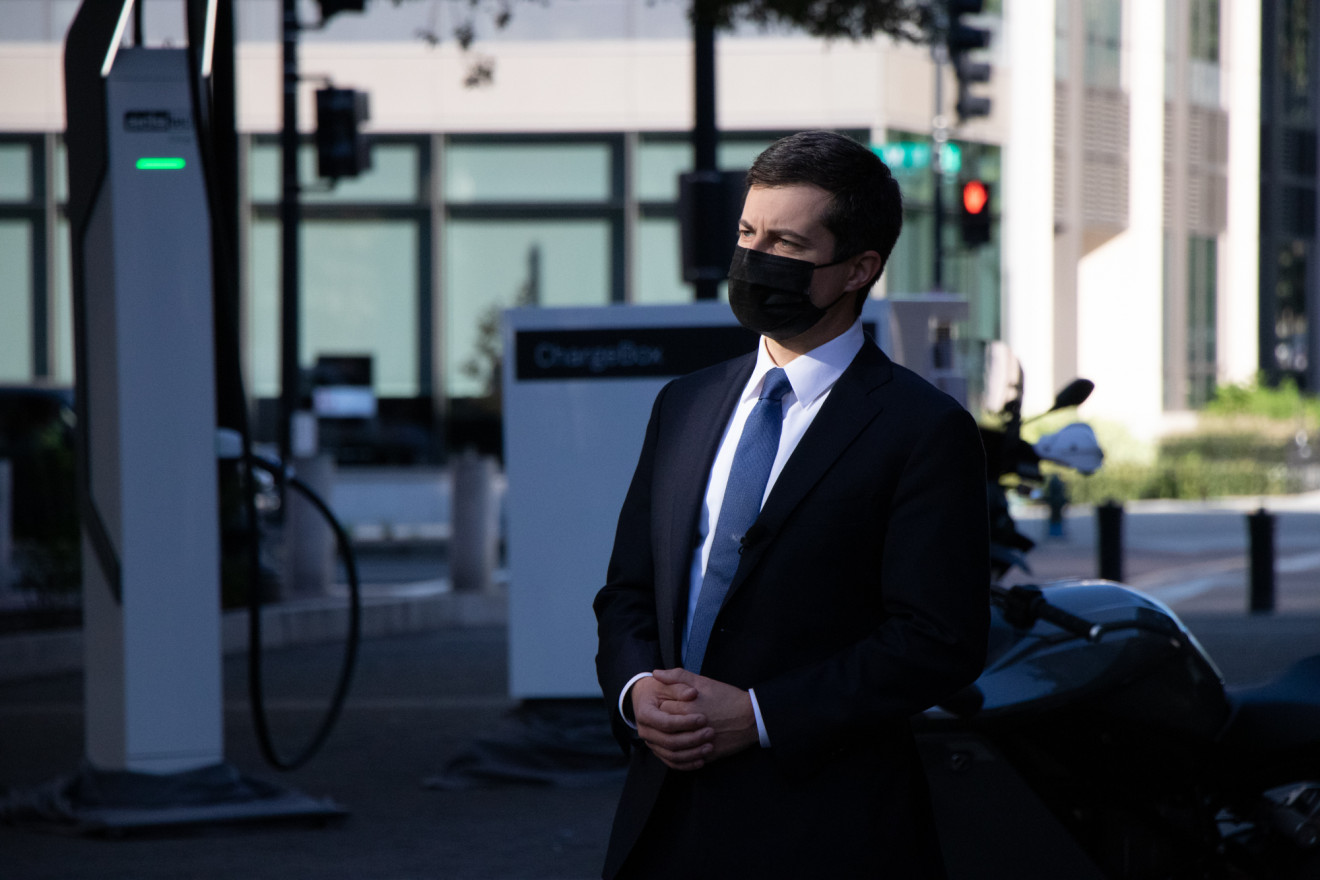 A man in a black suit, blue tie, and black mask stands with his hands interlaced with a EV charging station behind him.