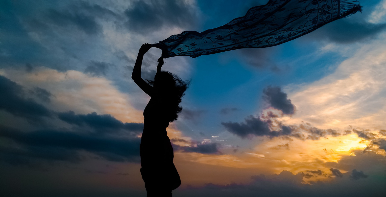 Silhouette of a woman waving a scarf in the sky against the backdrop of a cloudy sky at sunset.
