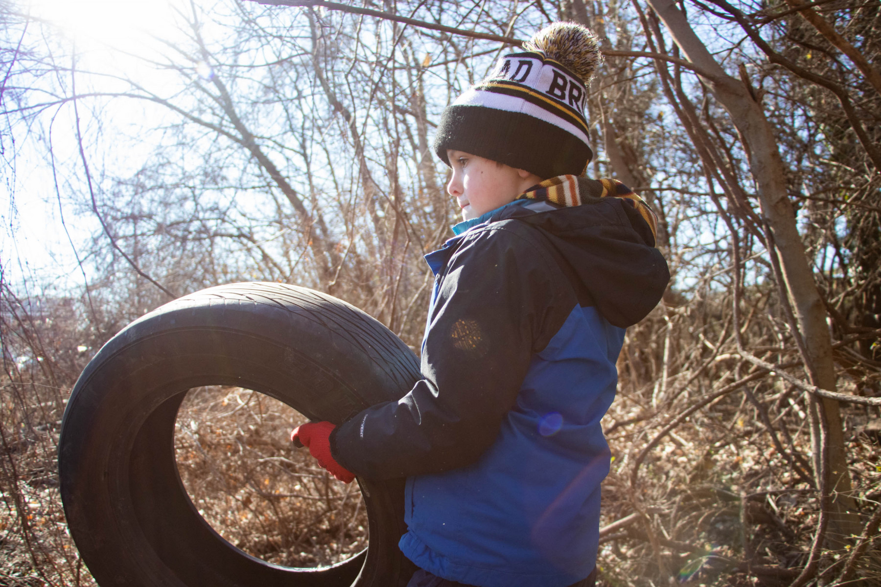 A young child in winter coat, hat, and gloves carries a car tire amidst a wooded background.