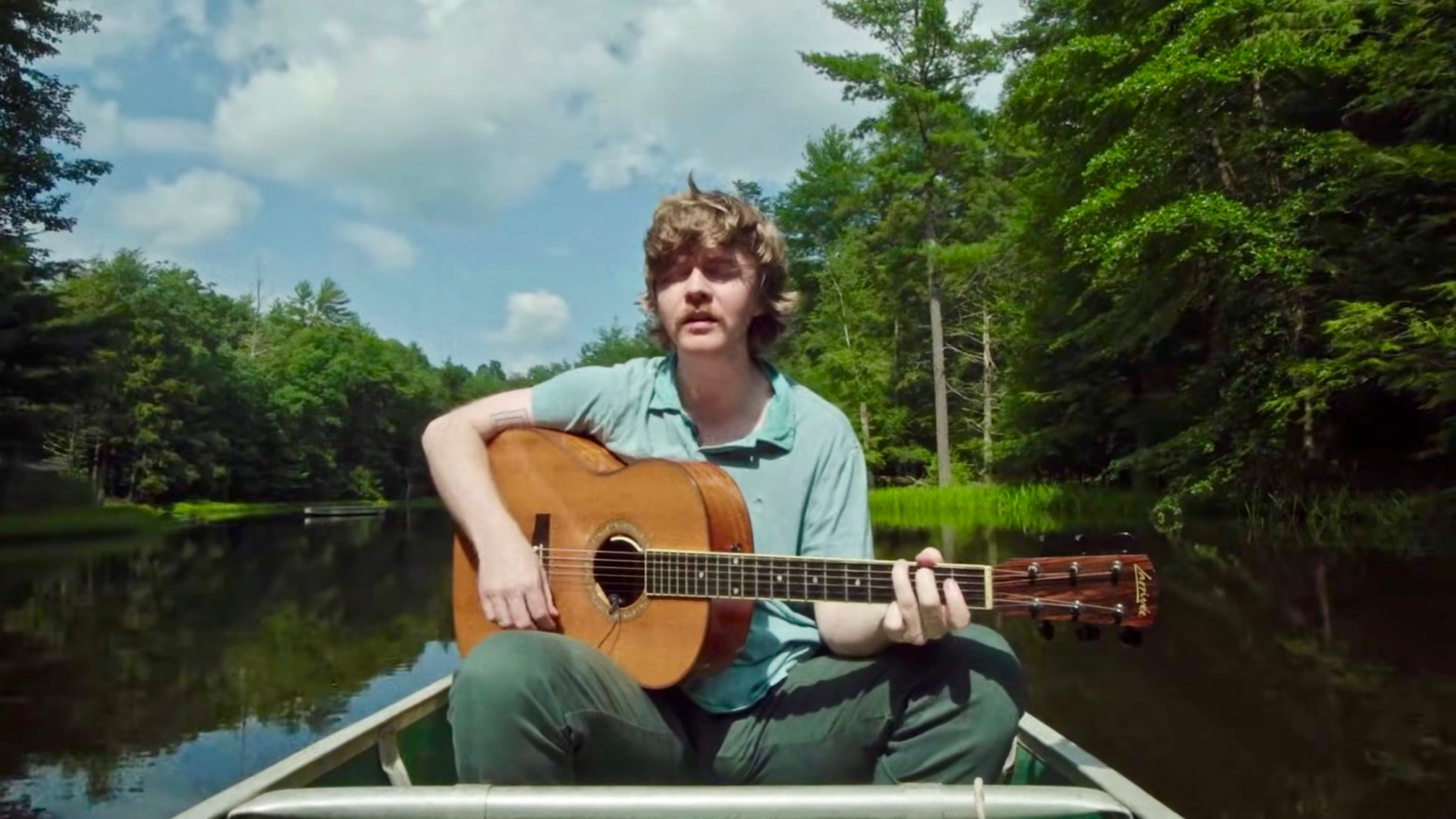 A man with dark blonde hair in a pale green polo and green pants plays guitar while in a canoe in a narrow body of water surrounded by lush green trees and a cloudy blue sky.