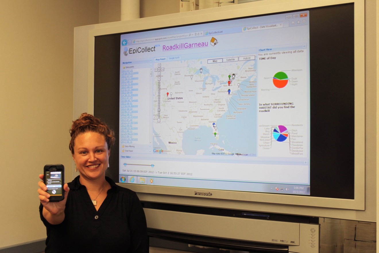 Woman in a black shirt smiles at the camera while holding out an iPhone and standing in front a large monitor showing a page titled "EpiCollect RoadkillGarneau" with a map of the continental U.S. and several charts.