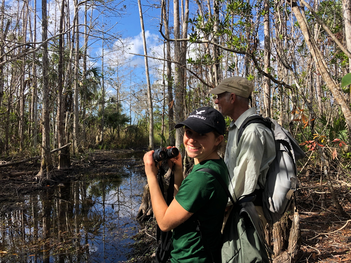 Young woman in a green shirt and black cap holds binoculars and stands next to a man in a neutral shirt and cap in a forested area next to a body of shallow water.