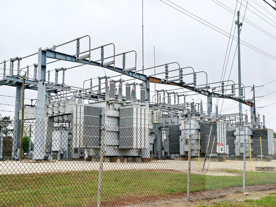 The gray machines of a transformer station sit behind a wire fence aside green grass.