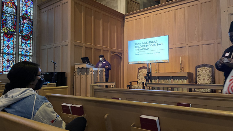 Person in mask sits in a church pew looks forward at a presentation. The presenter stands in front of a screen which reads "HOW INDIGENOUS PHILOSOPHIES CAN SAVE THE WORLD."