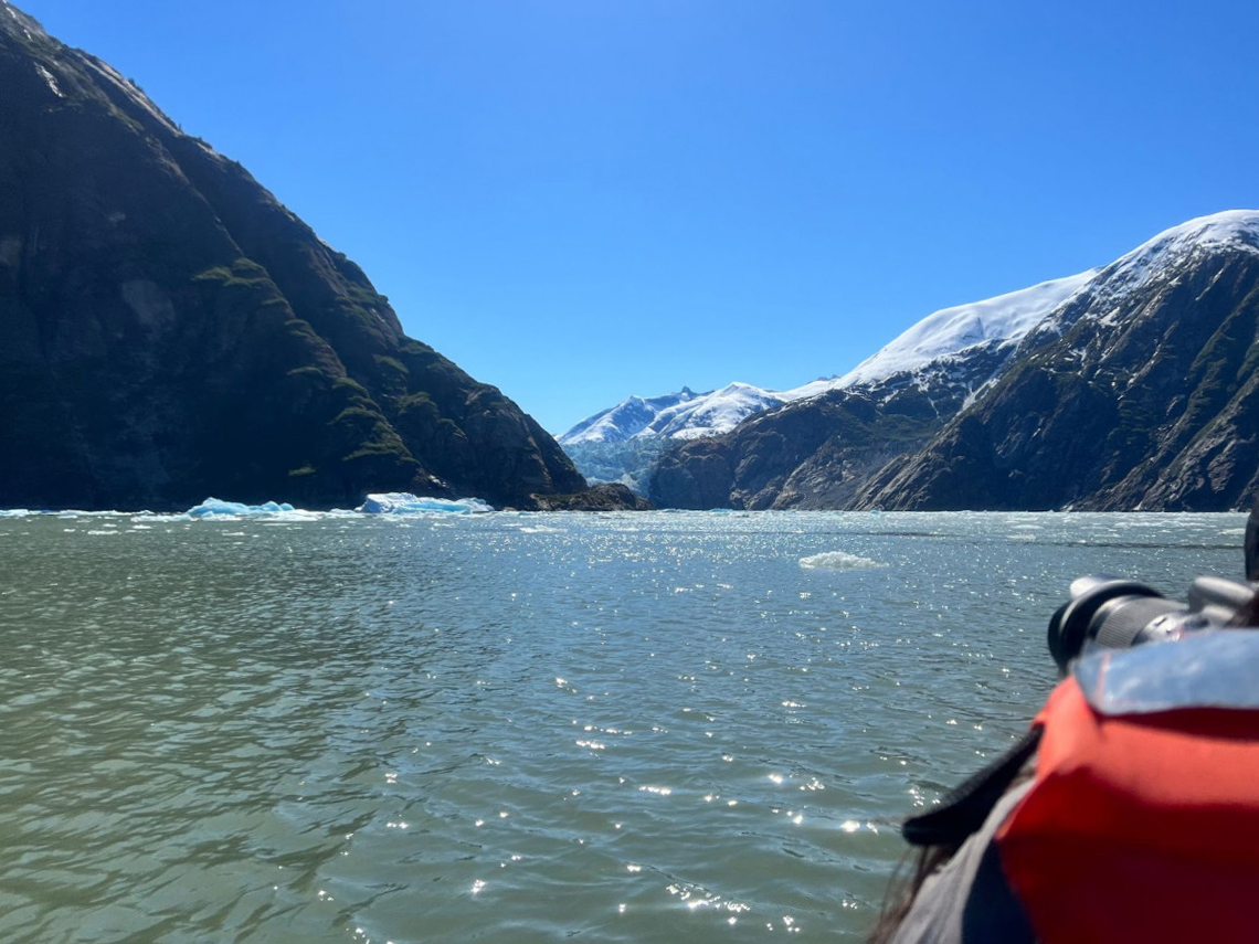 A bright blue sky stands behind towering mountains carved deep in the center by ancient glaciers. On the right a bright orange life jacket and camera lens are visible of a photographer capturing the scene aboard a small boat. Blue-green water is immediately in the foreground.
