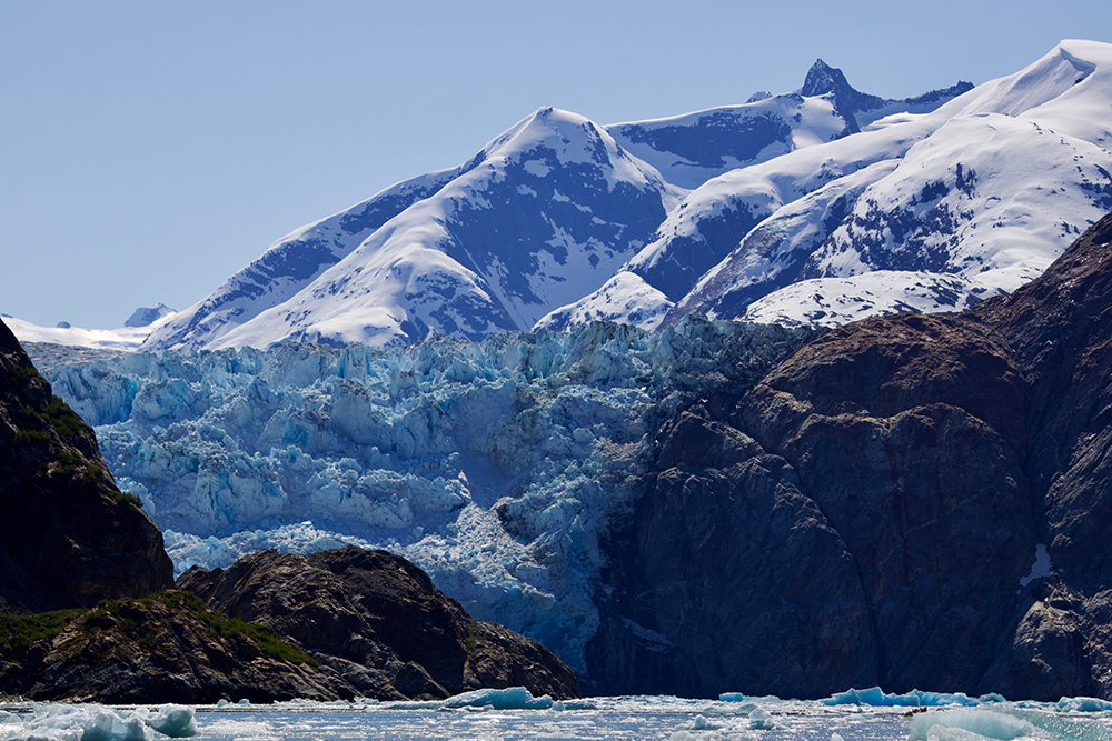 The face of a massive, icey blue glacier between two rocky landmasses.