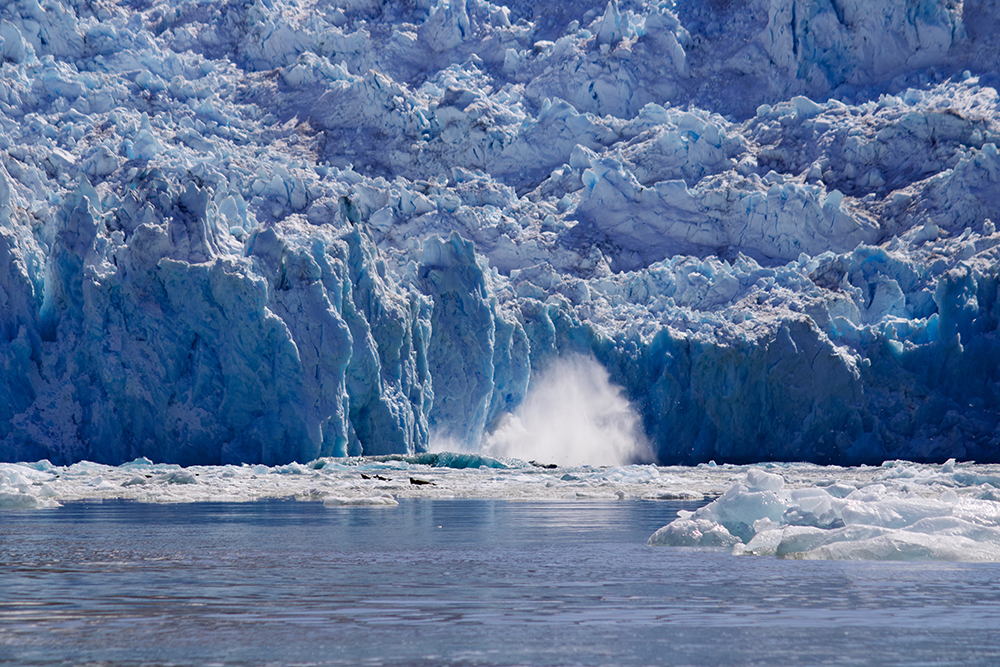 An upward splash of water can be seen surrounded by floating sea ice and a tall, vast glacier.