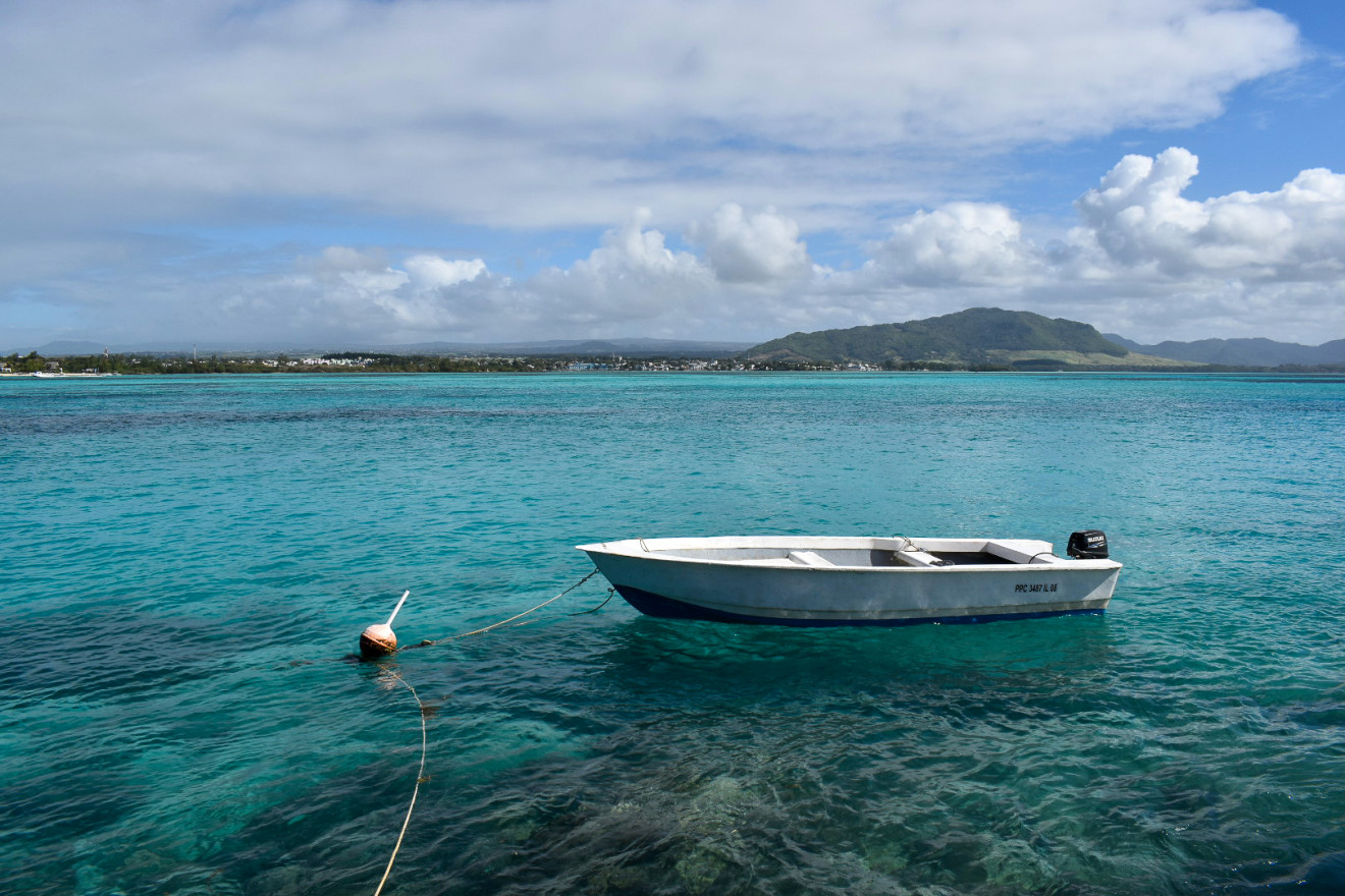 A small white boat rests buoyed on clear, blue-green waters under a cloudy blue sky with a gentle mountain in the distance.