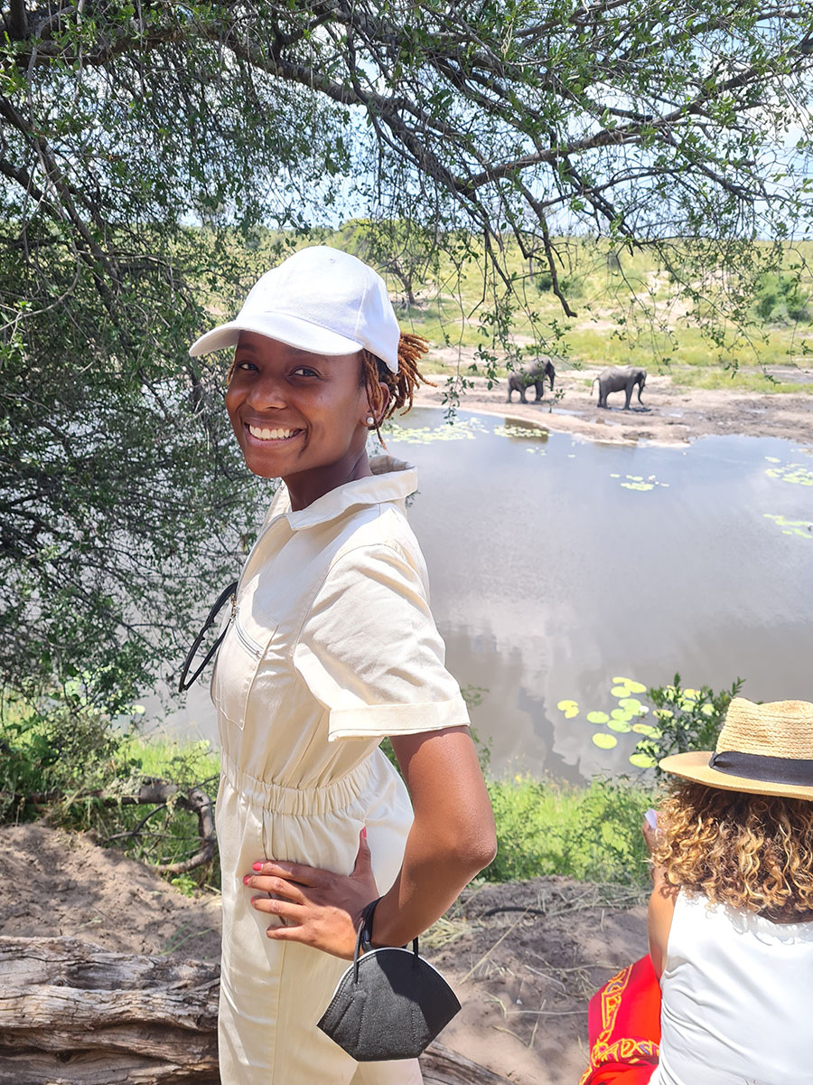A woman in a tan jumpsuit and a white baseball cap, with her hands on her hips and a black mask dangling from her left wrist, looks back at the camera smiling. Elephants are seen in the background near a body of water.