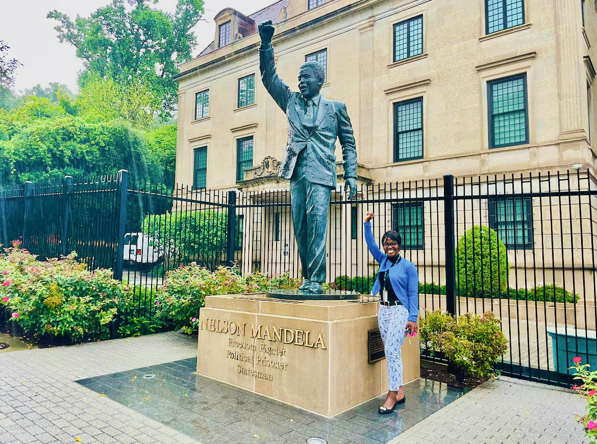 The author stands next to a statue of Nelson Mandela in front of a large, tan building. Both Beverly and Mandela hold up a raised fist. 