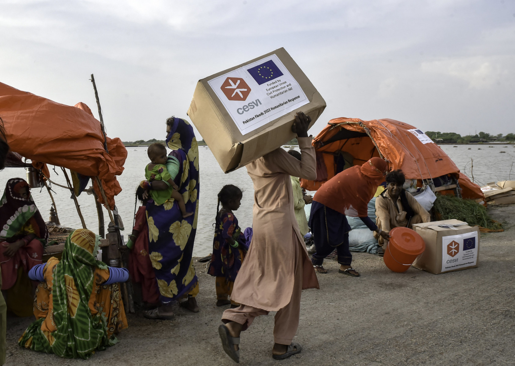 A person carries of a box of humanitarian aid passed a row of people who have been displaced from their homes. A plain of water stretches out into the distance behind them.