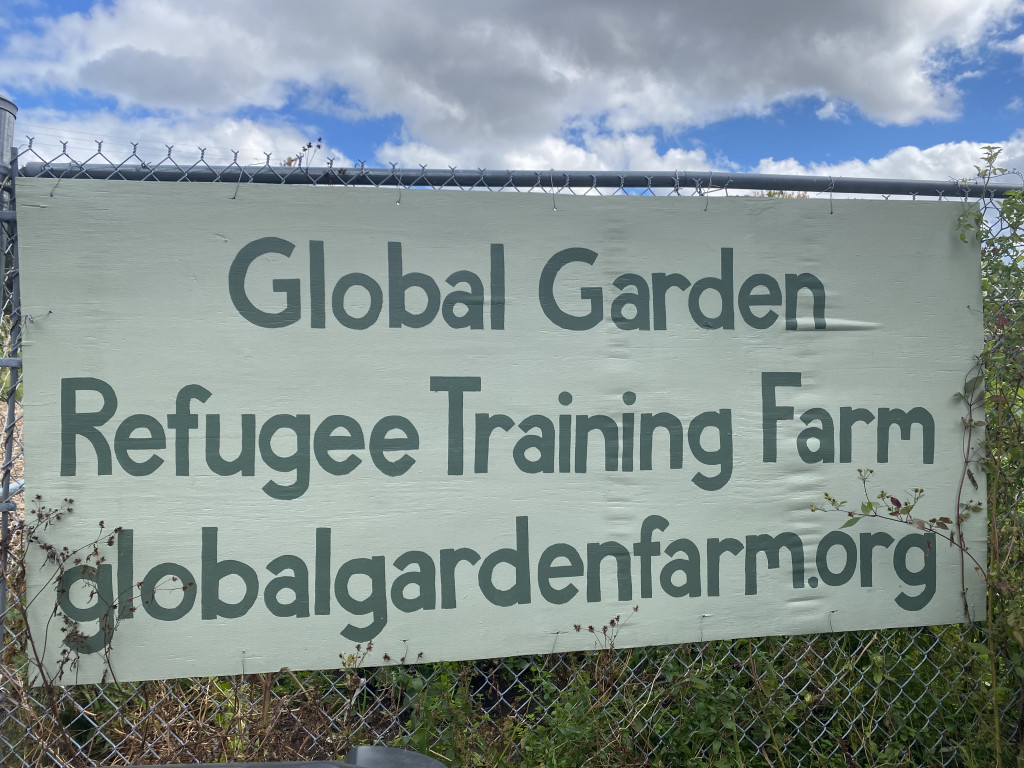 A sign hangs on a chain-link fence, reading "Global Garden Refugee Training Farm."