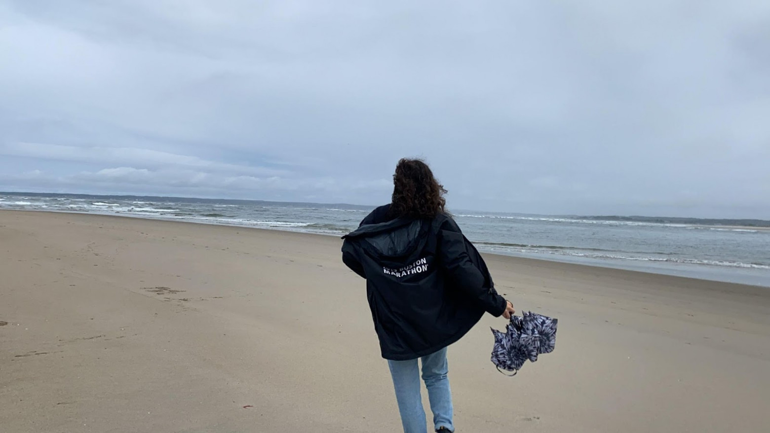A woman, seen from the back, is wearing light blue jeans and a black jacket carries a closed umbrella and is being blown back by the wind. She walks along a coastline's hard-packed sand on a cloudy, grey day.