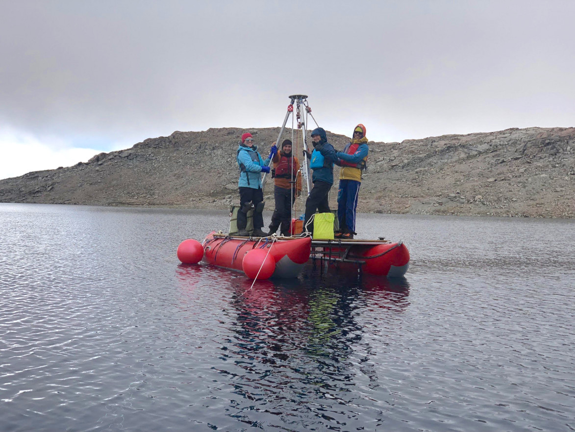 Four people stand on an inflatable raft in a body of water holding to a large piece of scientific equipment. 