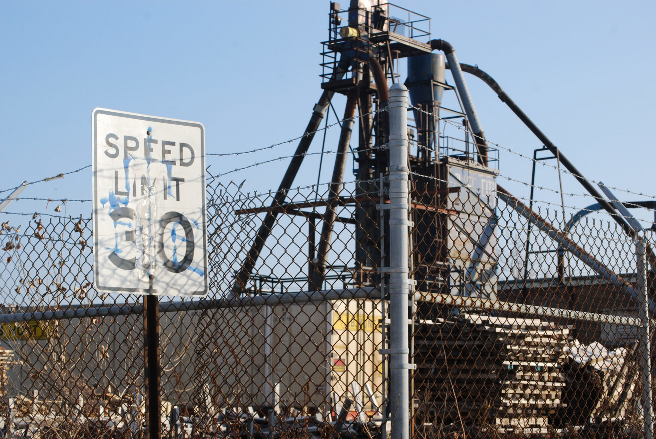 A factory tower looms behind a chainlink fence. A graffitied speed limit sign stands in the foreground.