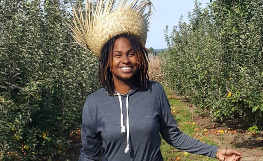 A woman stands between rows of cultivated plants, wearing a large straw hat and a grey hoodie, smiling at the camera.