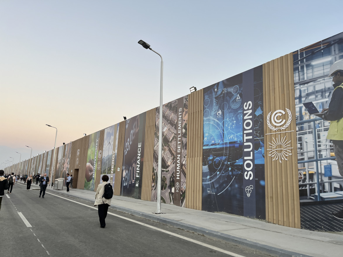A handful of people walk down a street next to a large wall that extends into the distance. Logos and advertisements for COP27 cover the entire length of the wall.