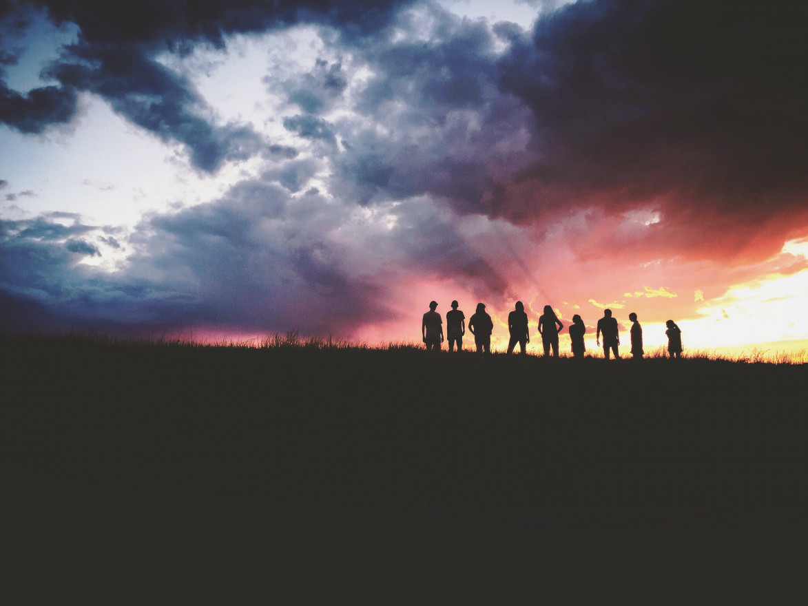 A group of young people stand silhouetted on a hillside with the sunset behind them.