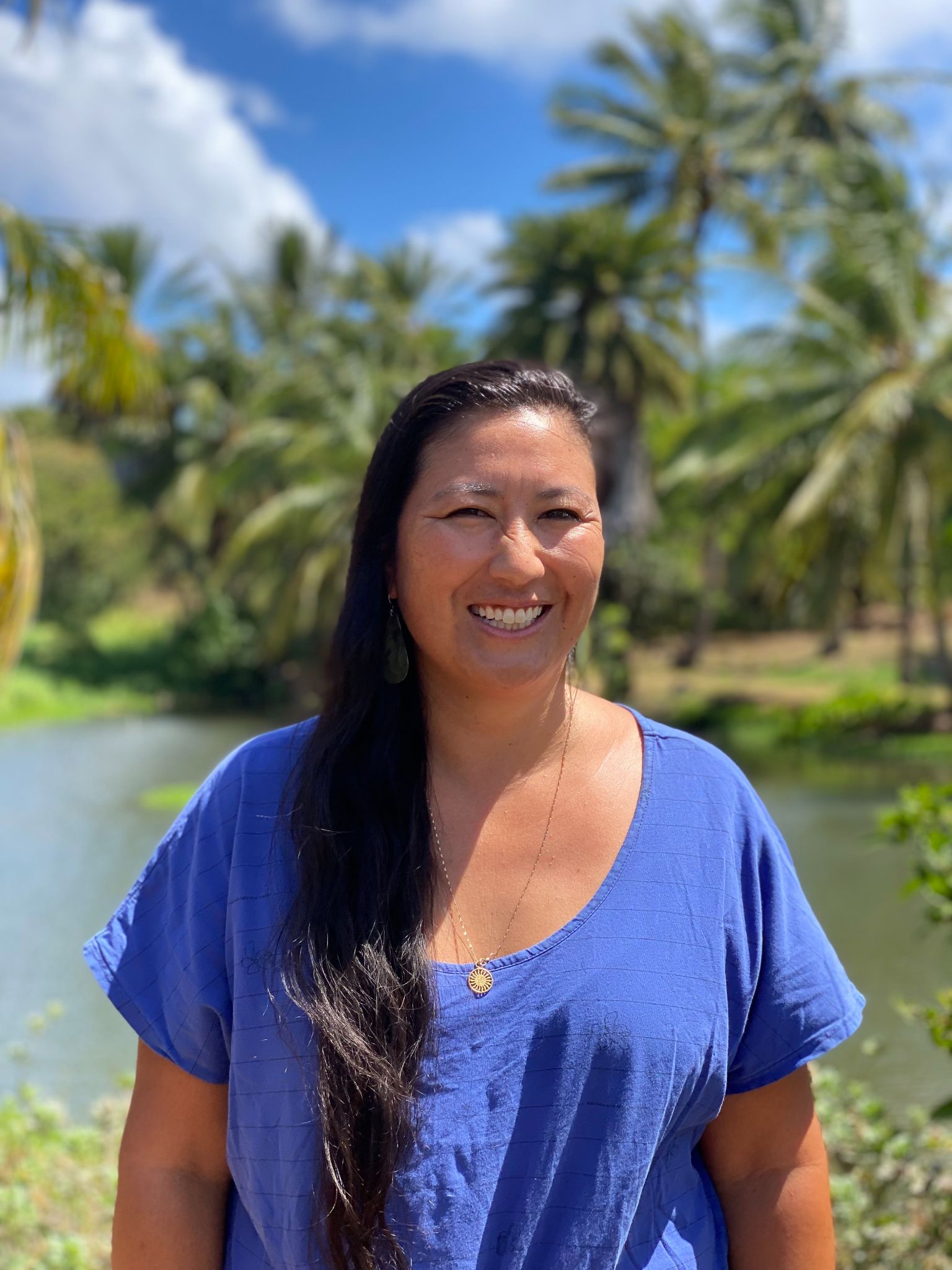 A woman with long, dark hair in a blue shirt smiles at the camera in front of a pond and palm trees.