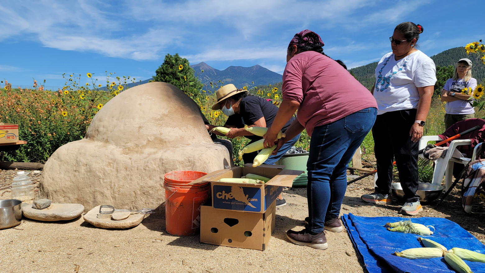 The women stand near a pueblo bread oven on a sunny day. Mountains are seen far in the distance.