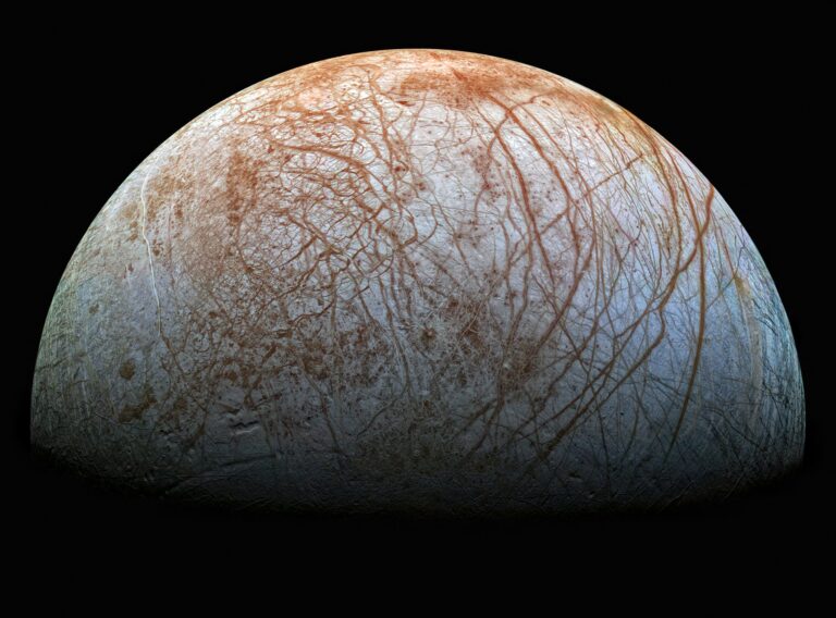 A half-lit sphere of Europa, which is a white with a blue tint, and covered in lines showing orange beneath.
