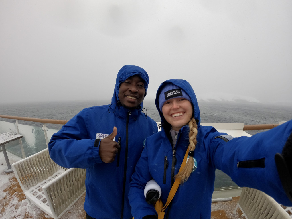 A woman and man in blue parkas smile on the snowy deck of a ship