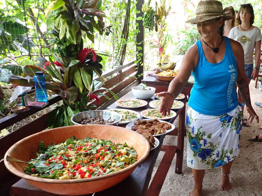 A person in a brimmed hat and a blue tank top stands near a table filled with large dishes of food in an outdoor, tropical setting.