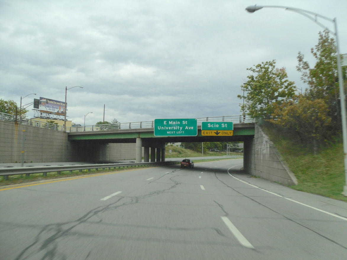 A portion of Rochester's Inner Loop taken from a driver's view. An upcoming bridge displays two different exit signs. No cars are on the expressway, except one in front of the camera.