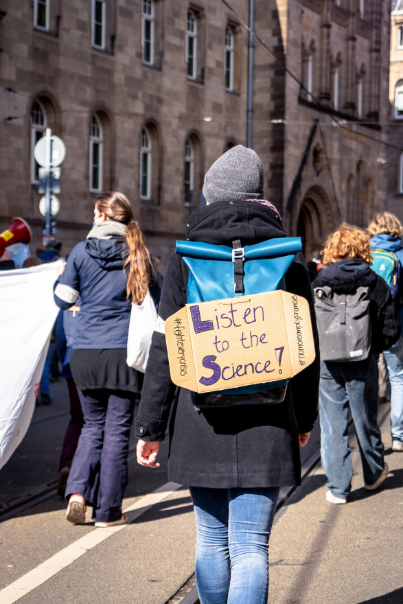 A person walks at a demonstration with a cardboard sign attached to their backpack, reading "Listen to the Science!"