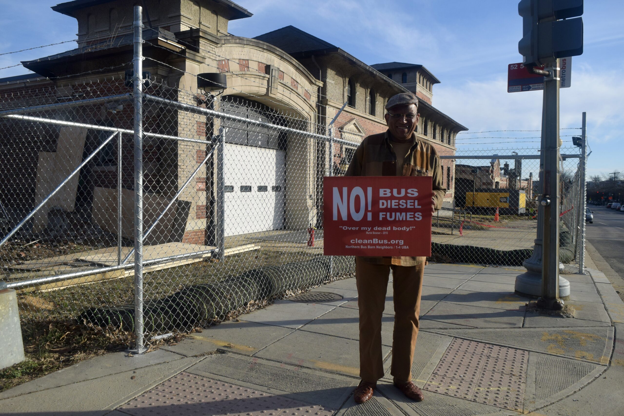 A man holds a red sign that says "NO! BUS DIESEL FUMES" outside of a brick building surrounded by wiring fencing.