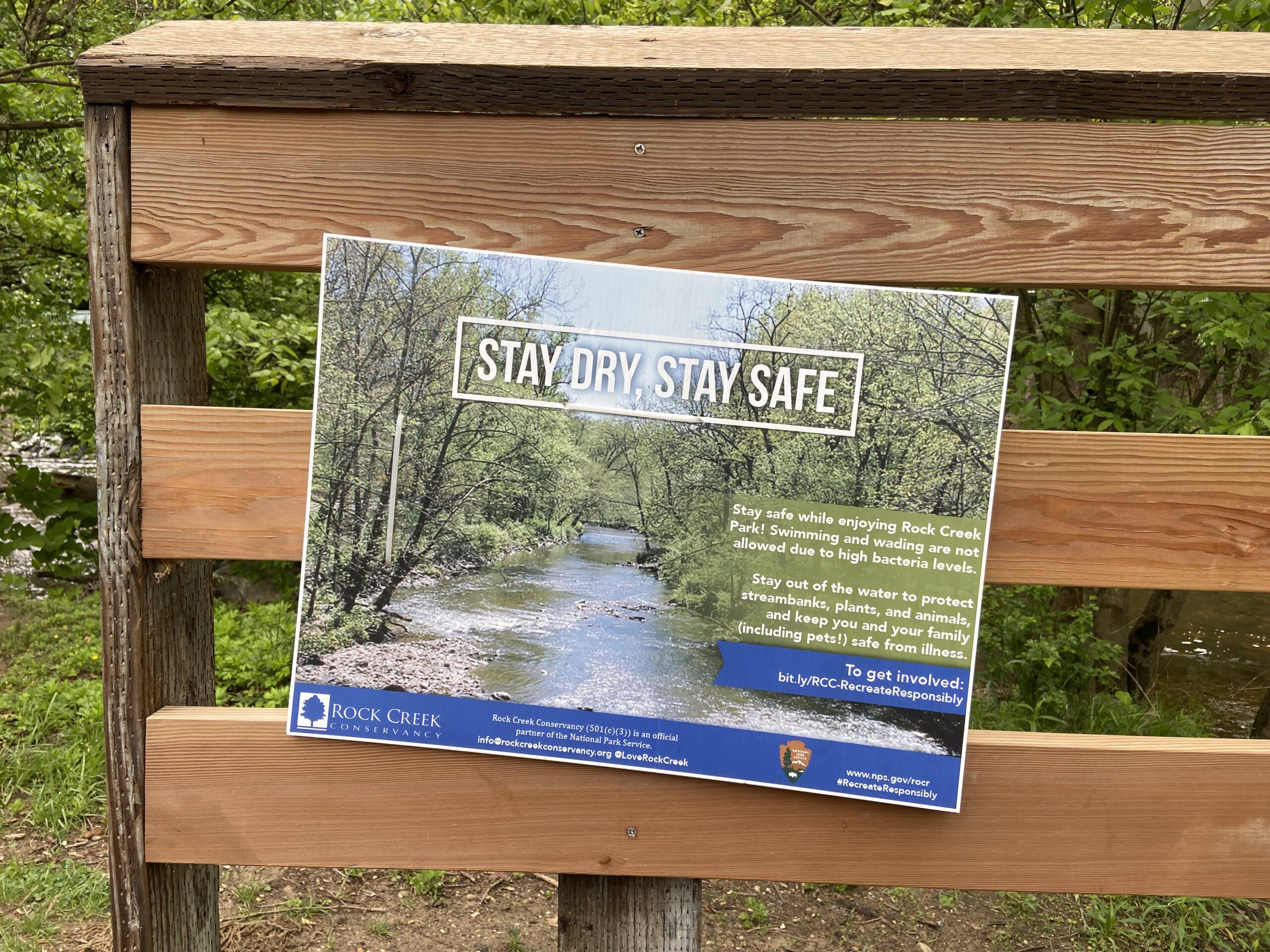 A sign on a wooden fence in a park reads "STAY DRY, STAY SAFE"