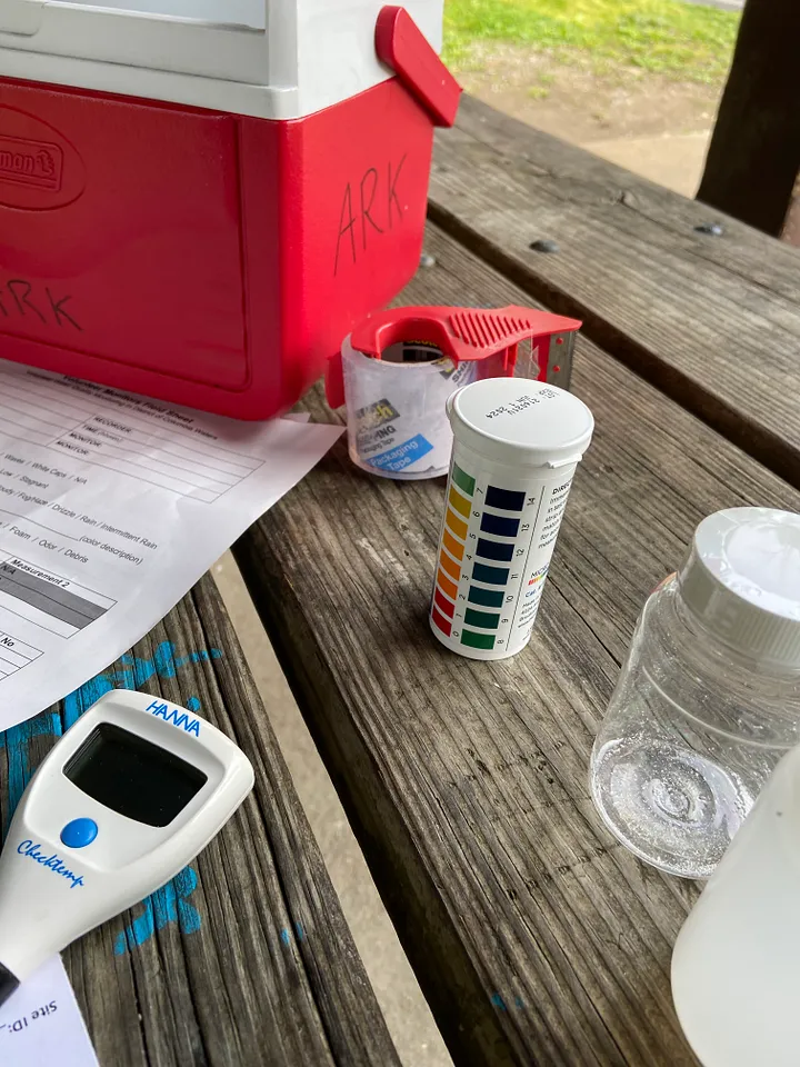 A small red cooler, roll of tape, thermometer, piece of paper, and small water sample jars sit on a picnic table.