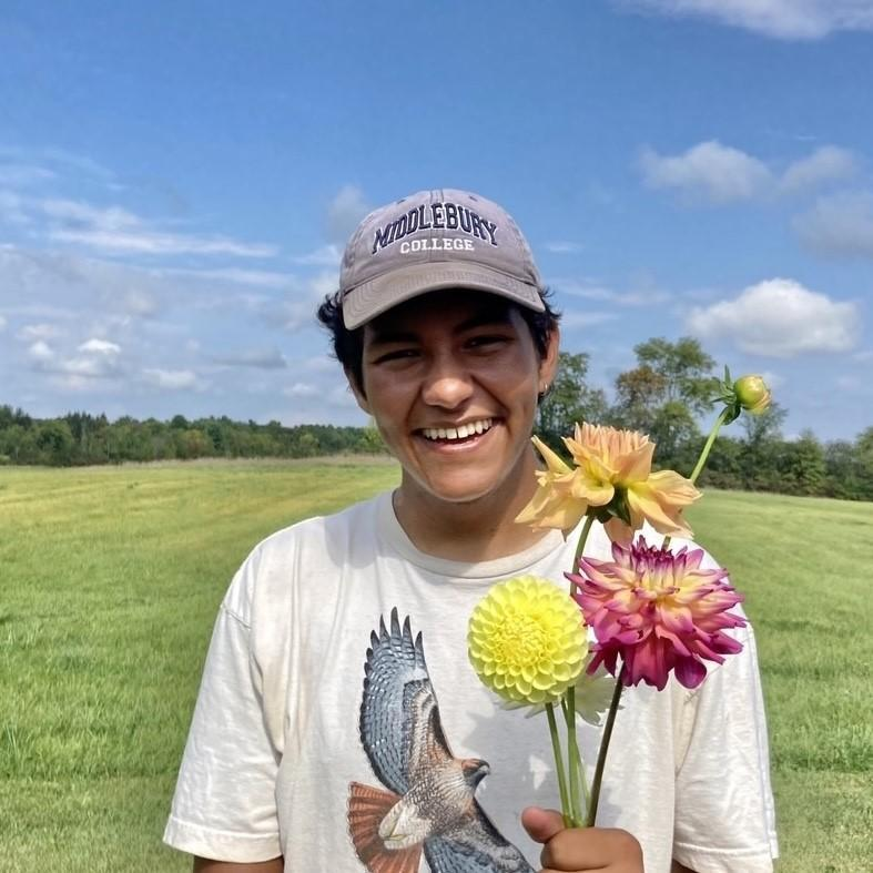 Andrés Oyaga, organizer for the Vermont Releaf Collect, stands in a field, holding several colorful flowers and smiling at the camera.