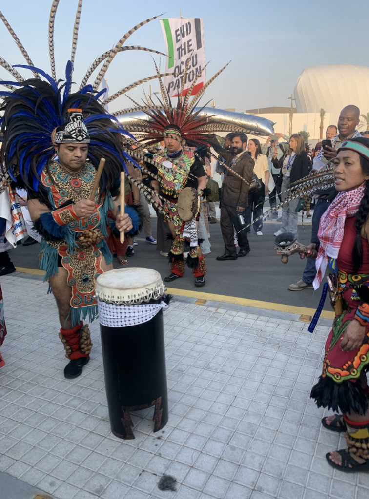 A person in Indigenous garb beats a drum outside COP28. 