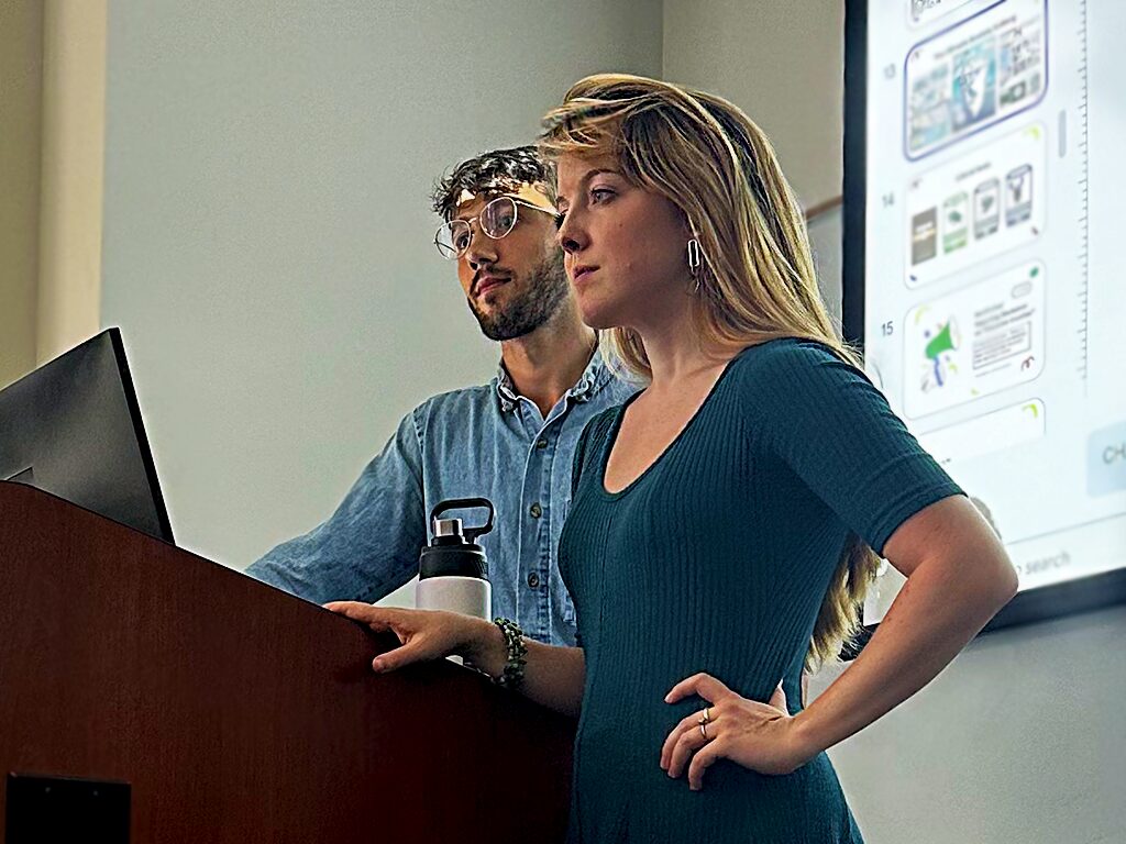 A man and a women stand in front of a lecture pedestal with a projector screen behind them.