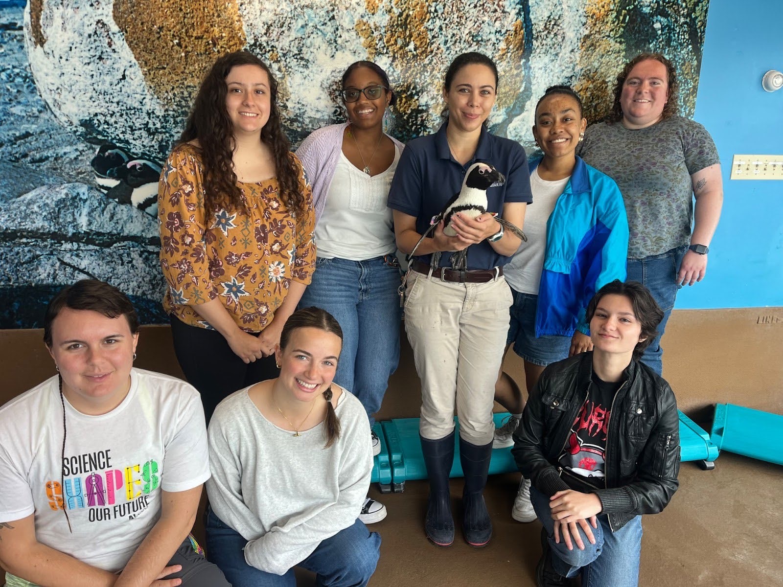 A group of students smile, facing the camera. The person in the center holds an African penguin.