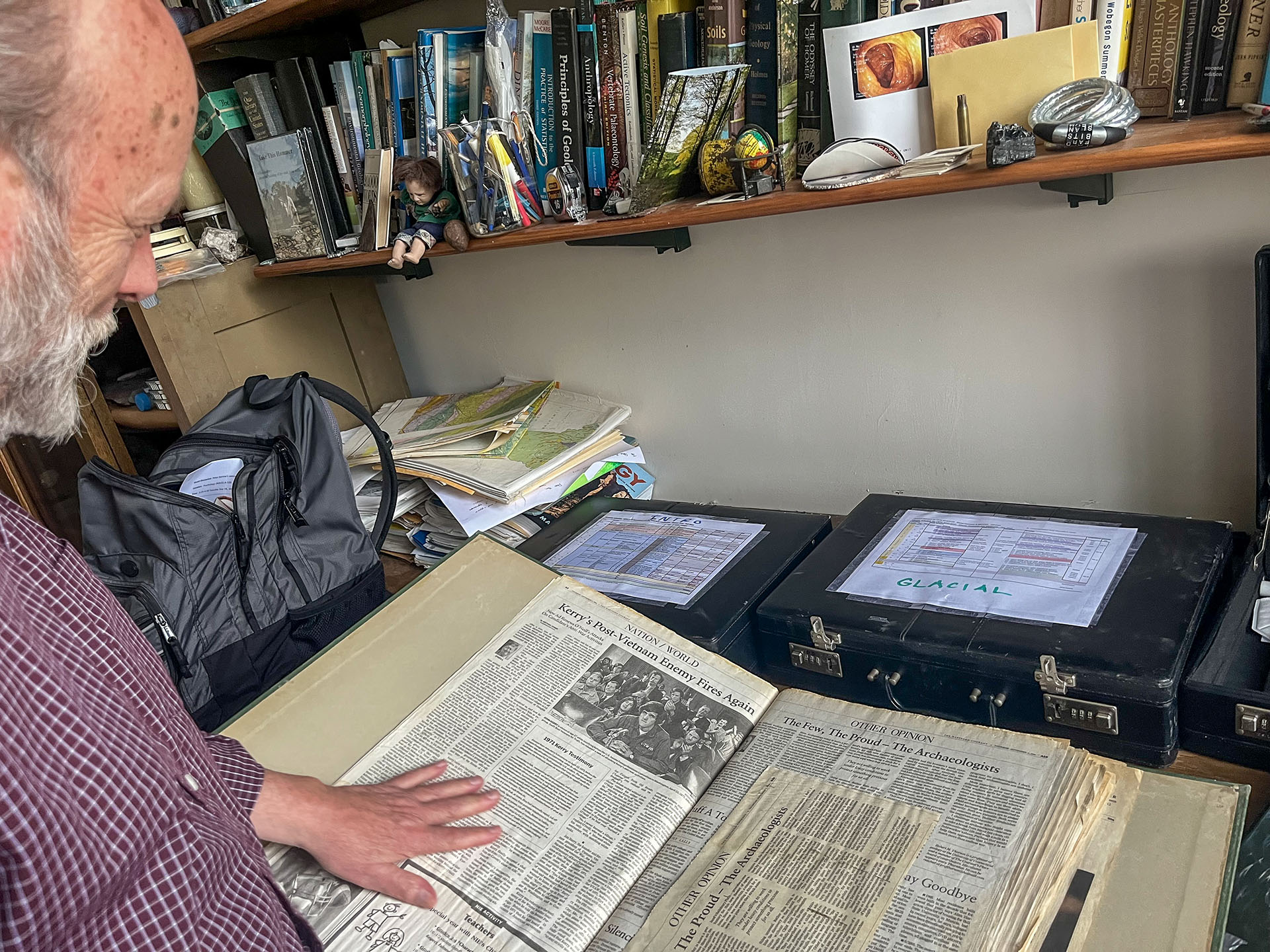 A smiling white male with a balding head and a white beard, and wearing a burgundy and white cross-hatched button down shirt, sits on a chair in his office. A large rock sits on a stool next to him and a brightly colored globe is in the foreground. A computer sits on the table behind him and a heavily laden bookshelf is in the background, with a window to the left of the frame.