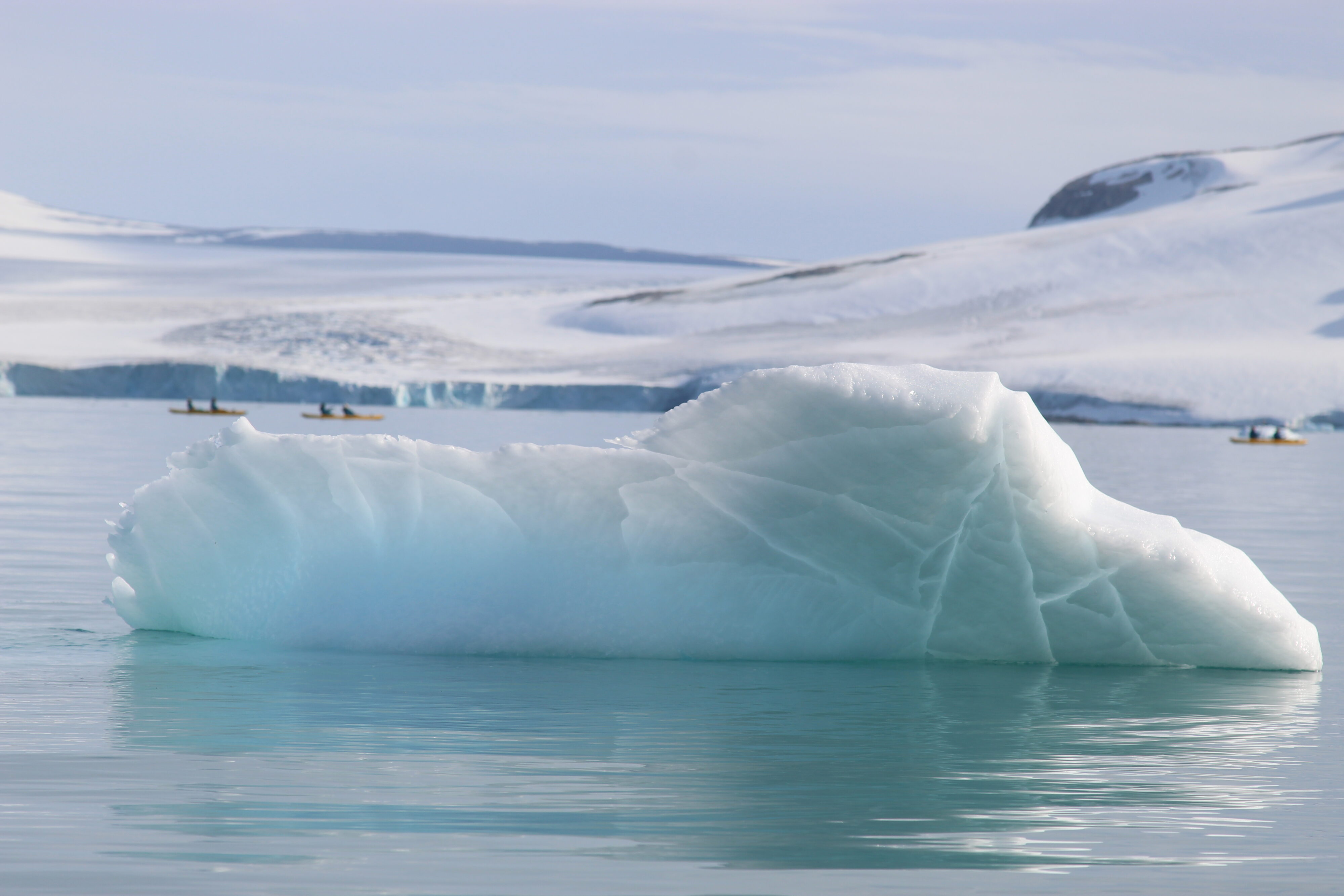 A light blue iceberg floats in teal blue waters. A massive glacier spans the background, and three yellow kayaks, each with two pasengers, dot the space between.